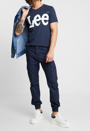 Tmavě modré bavlněné tričko s velkým bílým logem "Lee", doplněné tmavě modrými nákladními kalhotami a bílými teniskami s tmavě modrými detaily. Součástí je džínová bunda.