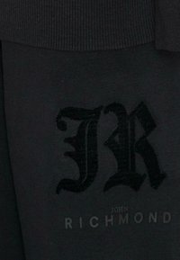 Zwarte stof met ribbels, met een reliëf "JR" logo en de tekst "JOHN RICHMOND" in een contrasterende afwerking.