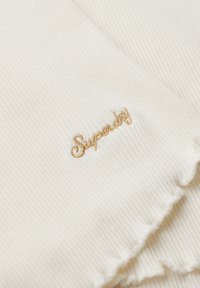 Hvid ribbet stof med en blød tekstur, der har "Superdry" broderet i guld. Kanterne har en frynsende finish.