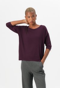 Fialový volný top s kulatým výstřihem a tříčtvrtečními rukávy. Modelka má na sobě šedé kalhoty na míru a stojí s jednou rukou na krku.