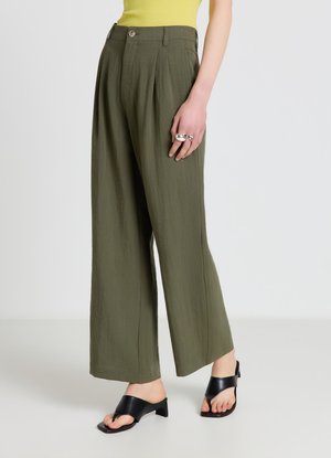 Femme portant un pantalon large vert olive, des sandales noires à talons ouverts, un haut jaune et des bagues argentées à la main droite.