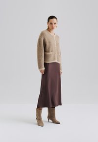 Beskjedent beige cardigan med tekstur, store knapper og lommer, iført over en brun satin midi-skirt og taupe ankelstøvler.