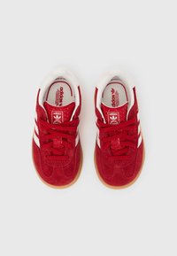 Červené semišové tenisky s bílými pruhy, kulatou špičkou, strukturovaným svrchním materiálem a gumovými podrážkami. Šněrování a polstrované límce zvyšují pohodlí.