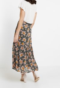 Frau in einem weißen kurzärmligen Oberteil, einem langen blumigen Rock mit pinken und orangen Blumen und beigen Blockabsatzsandalen, die seitlich steht.