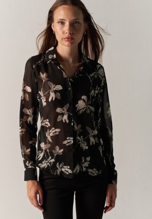 Mujer con una blusa negra transparente de manga larga y botones, con estampados florales blancos, de pie frente a un fondo claro y sencillo.