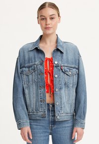 Giubbotto di jeans azzurro chiaro con una vestibilità rilassata, hardware argentato, due tasche sul petto e cuciture a contrasto, indossato sopra un top rosso.