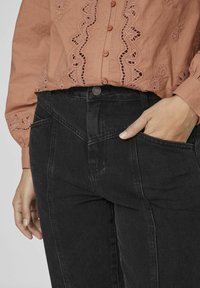 Blusa color óxido claro con detalles bordados y jeans negros de tiro alto con bolsillos en ángulo y cierre frontal de botones.
