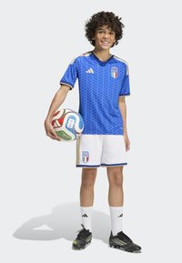 Blue soccer jersey with geometric pattern, gold accents, paired with white shorts. Black cleats and a multicolored soccer ball held.