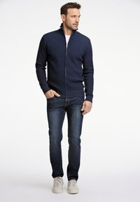 Marinblå zip-up tröja med en strukturerad design, kombinerad med mörk denimjeans och vita sneakers, stående mot en enfärgad bakgrund.