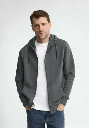 Hombre vistiendo una sudadera con capucha gris oscuro con cremallera sobre una camiseta blanca y jeans azules, de pie con las manos en los bolsillos frente a un fondo liso.