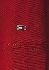 Tela roja con textura acanalada vertical, que presenta una etiqueta rectangular plateada con acentos rojos, blancos y marinos cosida cerca de la costura.