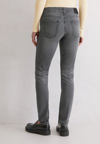 Personne portant un jean gris slim, des mocassins noirs et un haut en beige côtelé à manches longues, se tenant contre un mur texturé blanc uni.