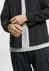 Camicia nera con bottoni e maniche lunghe, realizzata in tessuto liscio. Indossata sopra una t-shirt grigia chiara, con orlo dritto e vestibilità comoda.