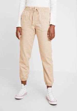 Personne portant un pantalon de jogging beige avec cordon de serrage et poches avant, ainsi que des baskets montantes blanches, debout devant un fond uni.