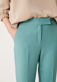Turkusowe spodnie z gładkiego materiału z płaskim przodem, bocznymi kieszeniami i dopasowanym krojem. Zestawione z beżową, miękko teksturowaną bluzką.