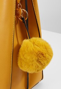 Bolso de cuero amarillo con acentos negros. Presenta un llavero de pom-pom amarillo esponjoso sujeto con un cordón negro. Superficie texturada.