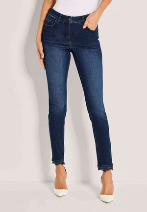 Woman wearing dark blue skinny jeans with frayed hems and white high-heeled shoes, standing against a plain light background.