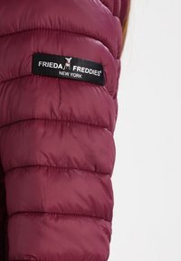 Burgundowa kurtka puchowa z przeszywanymi sekcjami oraz czarną metką z napisem "FRIEDA FREDDIES NEW YORK" w białym kolorze. Gładka tekstura.
