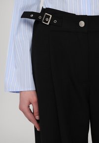 Zwarte broek met een getailleerde pasvorm, een brede tailleband, verstelbare riem met metalen details en zijzakken. Lichtblauw gestreepte blouse zichtbaar.