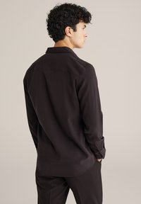 Črna dolga majica z dolgimi rokavi in klasičnim ovratnikom, gladka tkanina, gumbnice na zapestjih in ravno dno; kombinirana z ustreznimi temnimi hlačami.