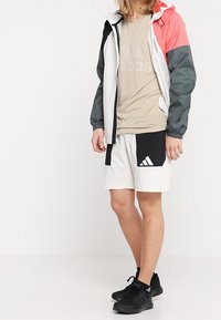 Mannelijke sportoutfit: lichtgewicht, kleurgeblokte jas met grijze, roze en zwarte secties; beige shirt; witte shorts met zwarte accenten; zwarte schoenen.