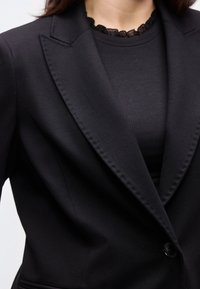 Γυναίκα που φοράει μαύρο σακάκι πάνω από μαύρη ριπτή μπλούζα με λαιμόκοψη διακοσμημένη με δαντέλα, εμφανίζεται από τους ώμους μέχρι το μέσο του κορμού.
