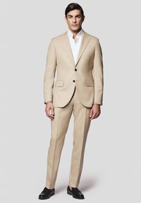 Beżowy garnitur wykonany z gładkiego materiału, z jednym zapięciem na guziki, dwoma przednimi kieszeniami i czarnymi guzikami, w połączeniu z czarnymi butami.