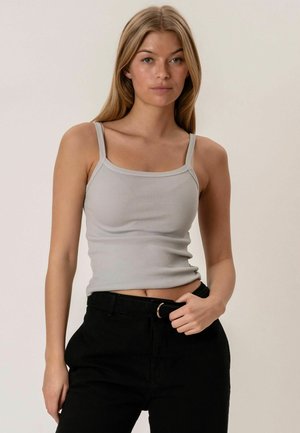 Jeune femme aux longs cheveux blonds portant un débardeur côtelé gris clair et un pantalon noir, debout devant un fond clair uni.