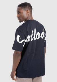 Černé tričko s krátkým rukávem vyrobené z bavlny, se velkým bílým kurzivním nápisem na zadní straně. Jednoduchý, uvolněný střih s kulatým výstřihem.