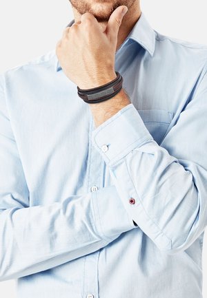 Pulsera multicapa en negro y gris llevada en la muñeca, con materiales texturizados. Camisa de color azul claro con botones, acentos de botones rojos en contraste.