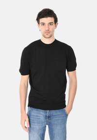 T-shirt nera a maniche corte realizzata in tessuto morbido, con scollatura tonda e una piccola tasca sul petto. Il modello indossa jeans blu strappati.