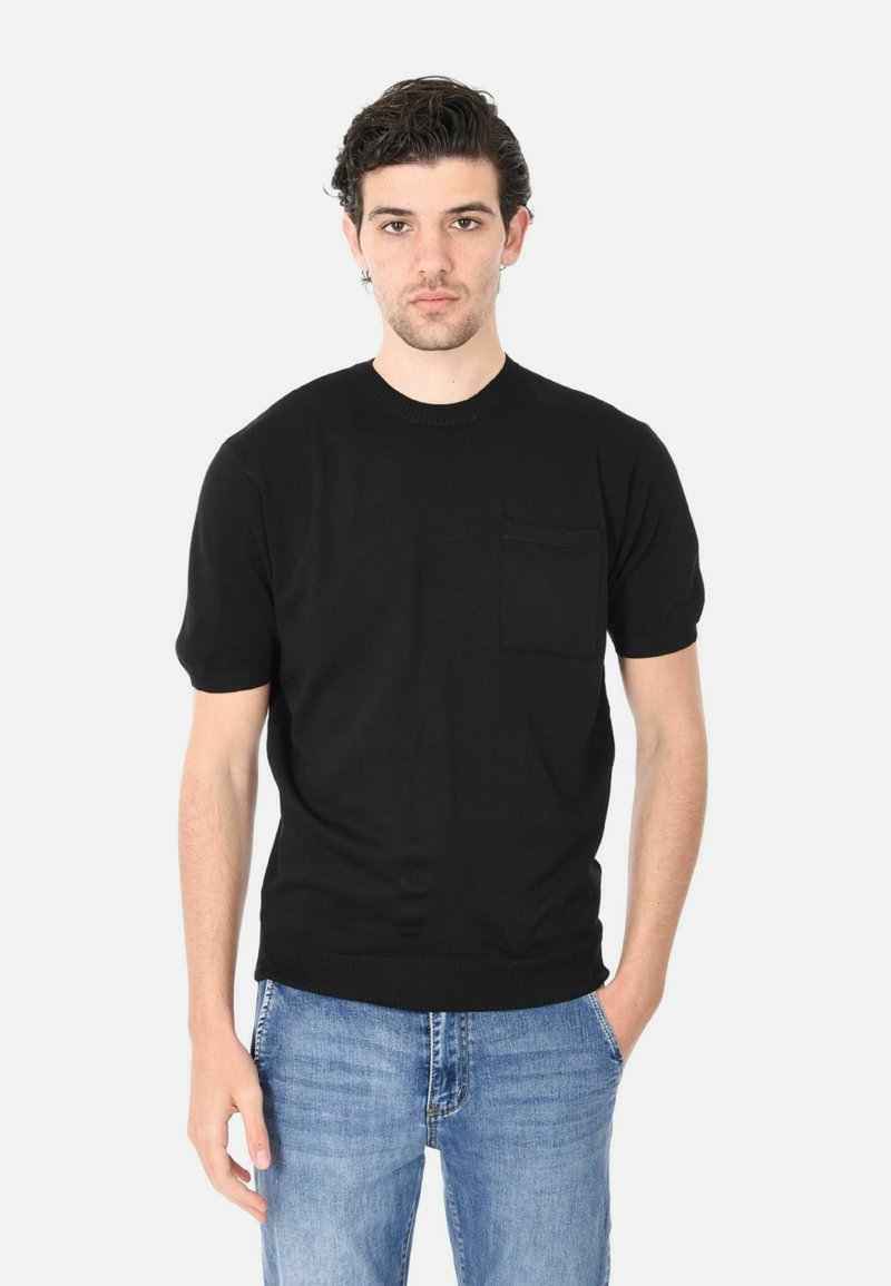 T-shirt nera a maniche corte realizzata in tessuto morbido, con scollatura tonda e una piccola tasca sul petto. Il modello indossa jeans blu strappati.