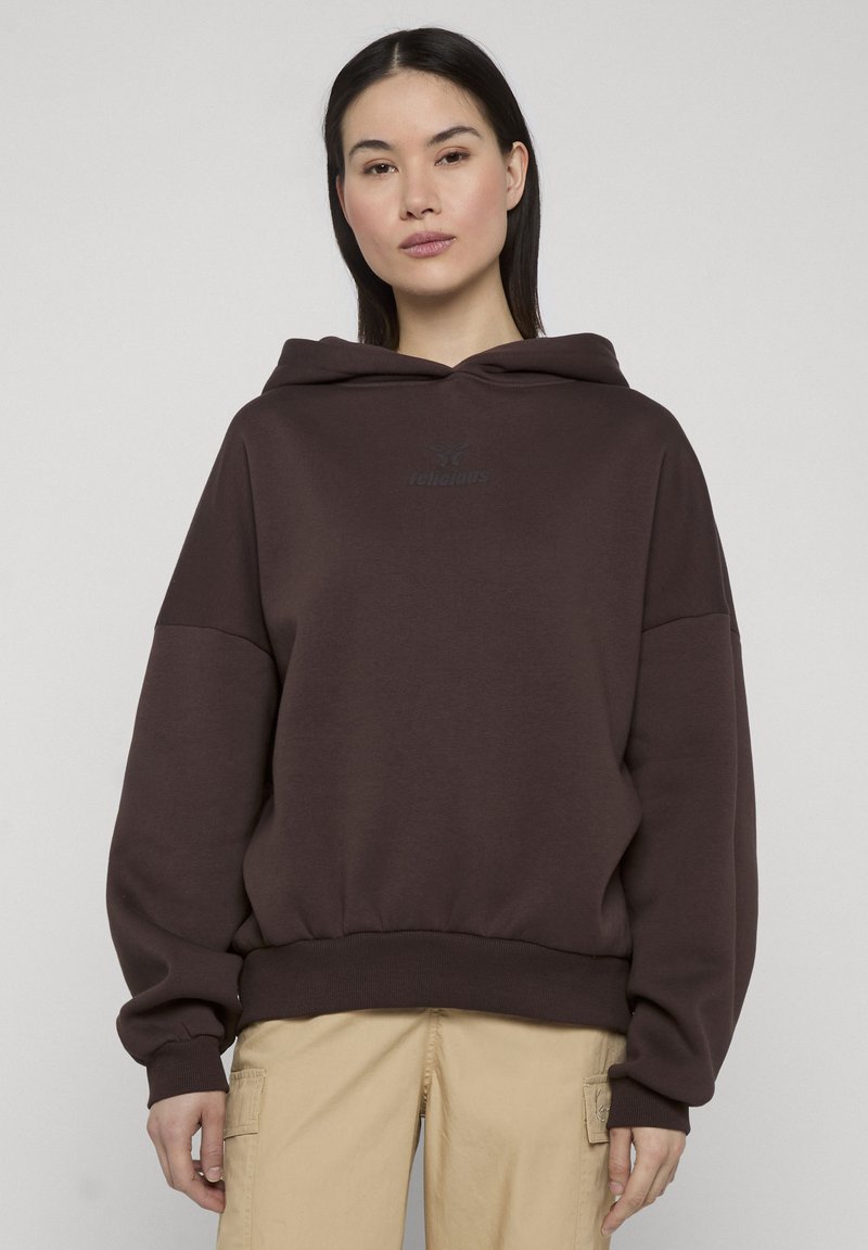Vrouw draagt een donkerbruine oversized hoodie en beige cargobroek, staand tegen een effen lichtgrijze achtergrond.