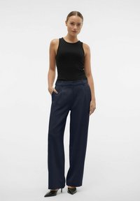 Zwarte mouwloze top en marineblauwe hoge taille broek met plooien. De broek heeft zijzakken en het model draagt zwarte puntige hakken.