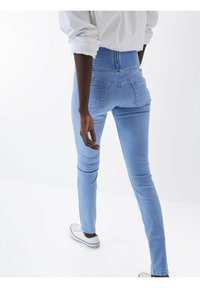 Jean skinny taille haute bleu clair en denim avec poches arrière discrètes et accents métalliques décoratifs. Design épuré avec couture minimale.