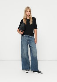 En blond kvinne står mot en hvit vegg iført en svart skjorte med knapper, vide blå jeans, svart-hvite joggesko og holder en svart clutch.