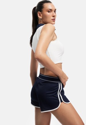 Jonge vrouw met paardenstaart, gekleed in een witte mouwloze cropped top en marineblauwe sportshorts met witte bies, poseert zijwaarts.