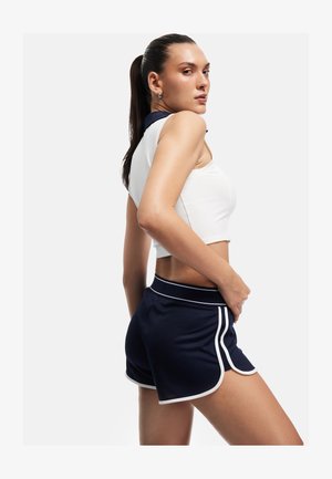 Jonge vrouw met paardenstaart, gekleed in een witte mouwloze cropped top en marineblauwe sportshorts met witte bies, poseert zijwaarts.