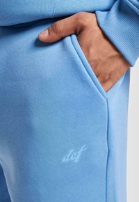 Helle blaue Jogginghosen mit einer glatten Textur, ausgestattet mit einer seitlichen Tasche und einem dezenten Logo, das auf dem Stoff gedruckt ist.
