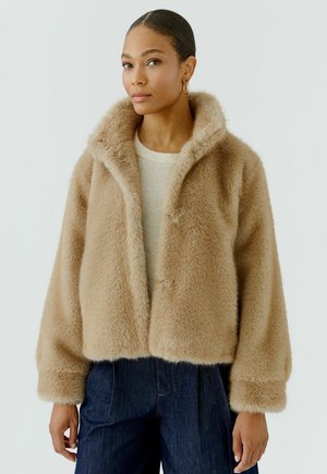 Vrouw draagt een beige jas van nepbont over een lichte trui en donkerblauwe broek, staand tegen een effen lichte achtergrond.