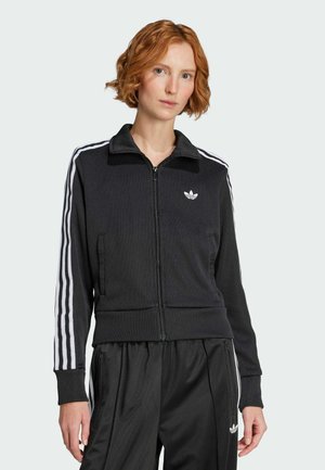 Rothaarige Frau trägt eine schwarze Adidas Trainingsjacke und -hose mit weißen Streifen und Logos, steht vor einfachem Hintergrund.