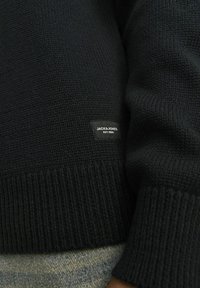 Fekete kötött pulóver, bordázott aljjal és mandzsettákkal. Egy kis szövetcímke látható, amelyen az áll: "JACK & JONES EST. 1990."
