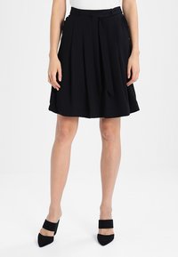 Jupe plissée noire avec une ceinture à nouer, en tissu lisse. Présente un design évasé et arrive au-dessus du genou. Associée à des talons noirs.