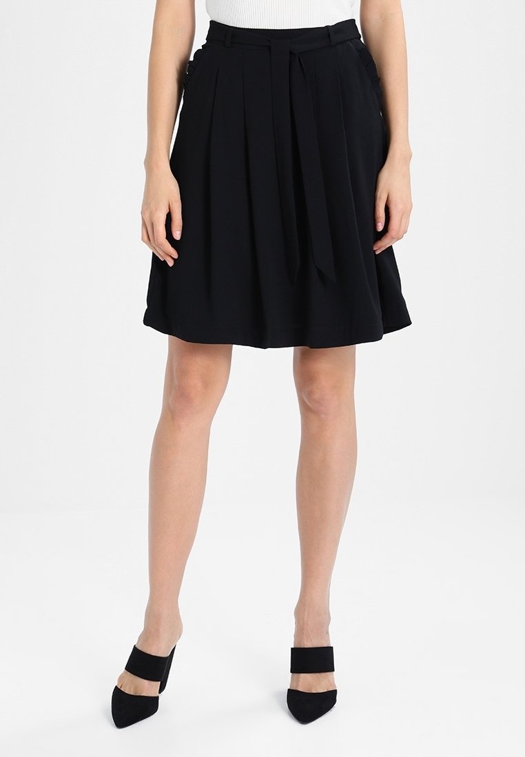 Jupe plissée noire avec une ceinture à nouer, en tissu lisse. Présente un design évasé et arrive au-dessus du genou. Associée à des talons noirs.