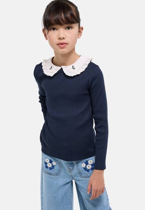 Top à manches longues côtelé bleu marine avec un col volant blanc ornée de broderies florales. Associé à un jean en denim clair mettant en valeur des patchs floraux.