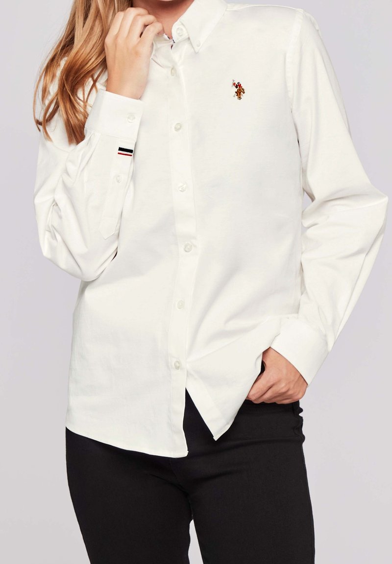 Femme portant une chemise blanche à manches longues boutonnée avec un petit logo brodé sur la poitrine et un pantalon noir, main près du col.