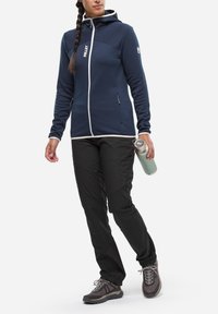 Granatowa polarowa kurtka z białymi akcentami, z zamkiem z przodu i kapturem. W połączeniu z czarnymi spodniami i szarymi butami, trzymając stalową butelkę na wodę.