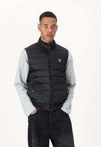 Black padded vest with high collar, front zipper, and two side pockets. Features a small logo patch and is worn over a gray long-sleeve shirt.