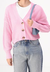 Person som bär en rosa tjock stickad kofta med stora knappar, vit skjorta, blåjeans med strassmönster, och håller en liten blå axelväska.