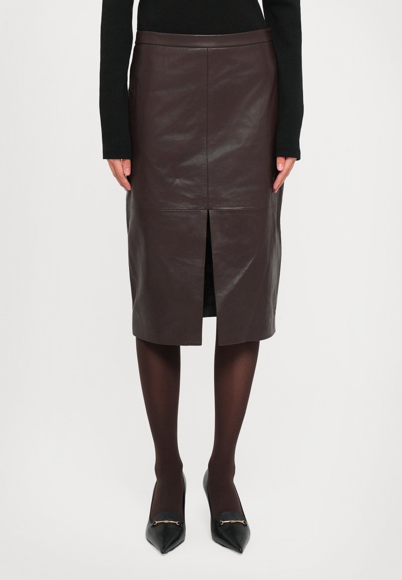 Woman wearing a dark brown leather knee-length skirt with a front slit, black tights, black pointed-toe heels, and a black long-sleeve top.